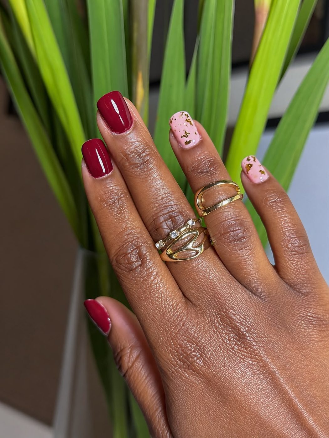 Burgundy and pale-pink manicure with gold foil photographed beside green leaves